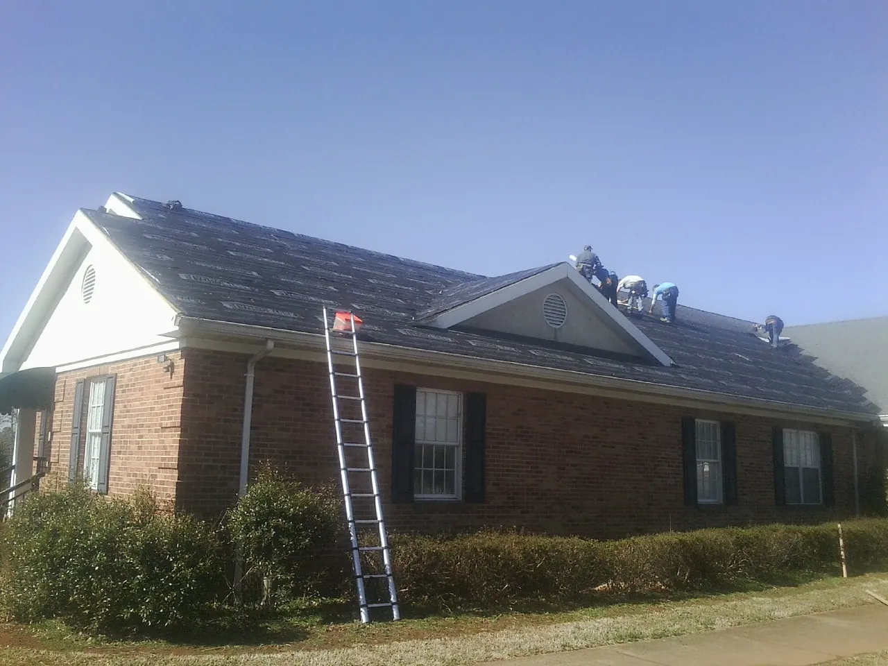 Crew tearing shingles off a brick ranch roof with a ladder set — Metal Roof Repair work by our crew in Bryan
