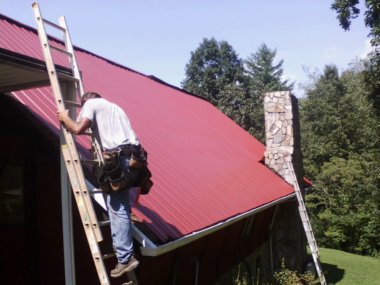 Roofer on a ladder beside a red metal roof and stone chimney — Soffit & Fascia Repair by Roofing Brothers in Bryan